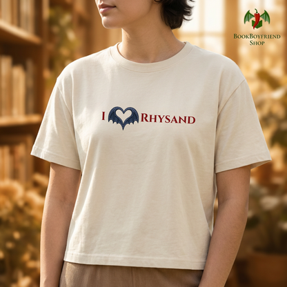 Person wearing a beige t-shirt with 'I ❤️ RHYSAND' design in front of a blurred bookshelf.